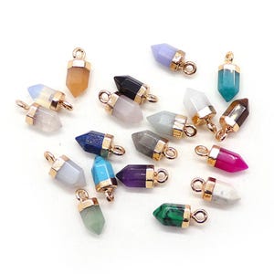 May include: A collection of small, gemstone pendants in various colors, including pink, green, blue, and purple. Each pendant has a gold-colored metal cap and loop for attaching to a chain or other jewelry component. The pendants are arranged on a white surface.