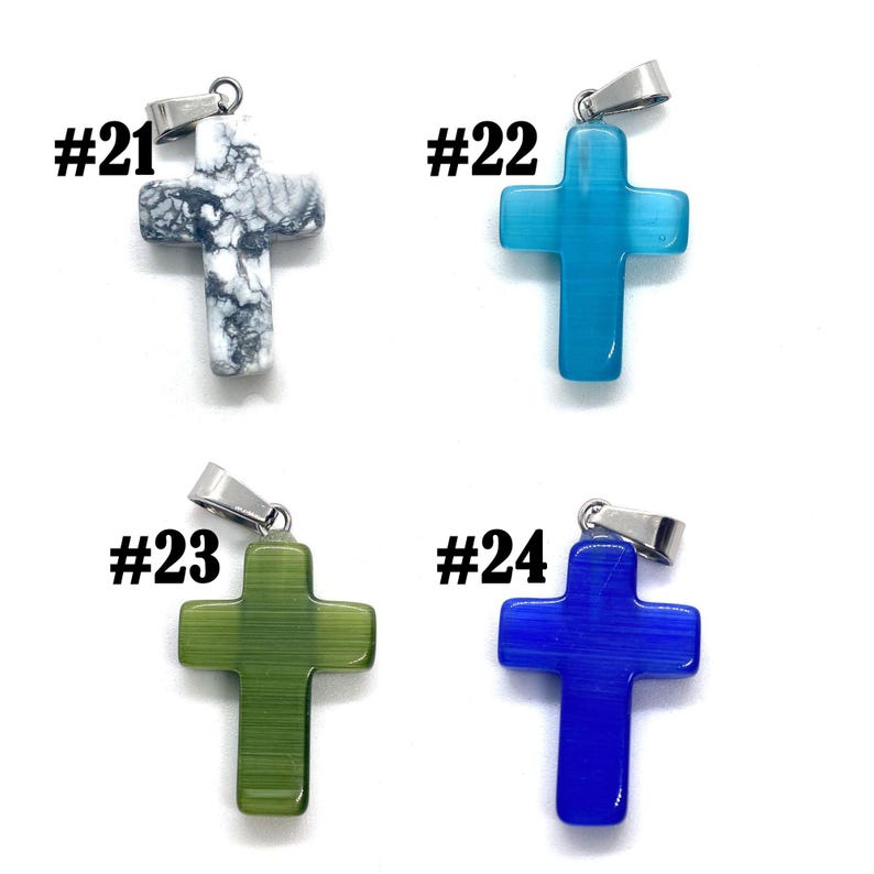 May include: Four cross pendants in various colors: white and gray, light blue, green, and dark blue. Each cross has a silver-colored bail and the numbers #21, #22, #23, and #24 are printed above each pendant. The crosses are made of a smooth, possibly stone-like material.