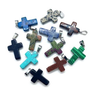 May include: A collection of gemstone cross pendants in various colors, including blue, green, brown, and black. Each cross has a silver-tone bail for attaching to a chain. The crosses are displayed on a white background.