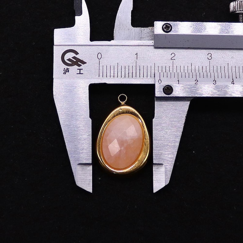 May include: A peach-colored, faceted gemstone pendant with a gold-colored bezel, measured with a metal caliper. The pendant has a small loop at the top for hanging. The caliper shows measurements in inches.