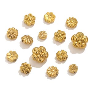 以下が含まれることがあります： さまざまなサイズの金色の花型のビーズのコレクション。ビーズには、詳細な花びらのデザインと、糸を通すための小さな穴があります。ビーズは白い表面に散らばっており、ジュエリー作りに視覚的に魅力的な配置になっています。