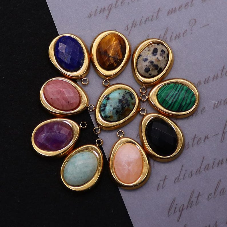 May include: A collection of oval-shaped gemstone pendants in gold-colored settings. The gemstones vary in color and pattern, including blue, brown, speckled, pink, turquoise, green, purple, black, and peach. Each pendant has a small loop for attaching to a chain or other jewelry component.