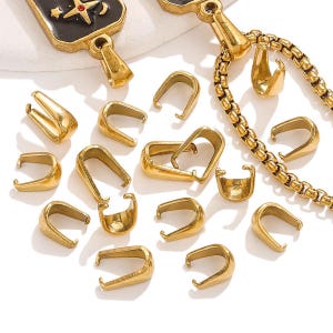 Puede incluir: Una colección de hallazgos de joyería dorados, que incluyen colgantes y componentes de cadena, dispuestos sobre una superficie blanca. Los colgantes son de varios tamaños y formas, con un diseño curvo para sujetar colgantes o dijes. También se ve una cadena dorada.