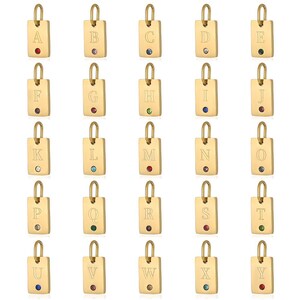 Może przedstawiać: Kolekcja prostokątnych wisiorków w kolorze złotym, każdy z wygrawerowaną literą alfabetu od A do Y. Każdy wisiorek posiada mały, kolorowy kamień szlachetny. Wisiorki są przeznaczone do biżuterii.