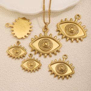 Peut inclure: Bijoux dorés, dont un collier et plusieurs pendentifs. Les pendentifs présentent un motif d'œil avec des détails complexes. Le collier a une chaîne délicate. Les pièces sont disposées sur une surface blanche.