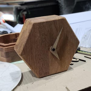Könnte beinhalten: Eine Holzuhr mit sechseckigem Zifferblatt. Die Uhrzeiger sind hell und zeigen ungefähr 10:10 Uhr an. Die Uhr ist aus warm getöntem Holz gefertigt. Im Hintergrund ist eine Holzkiste zu sehen.