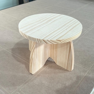 May include: A small, unfinished wooden stool with a round top and three curved legs. The natural wood grain is visible on the surface. The stool is approximately 30 cm in diameter and 25 cm tall.