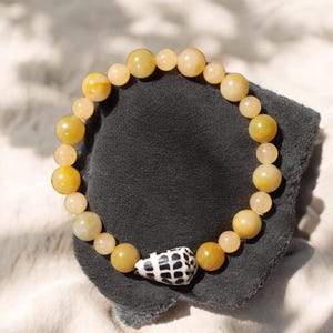May include: A beaded bracelet featuring yellow and cream-colored round beads. A white and black seashell charm is centered on the bracelet. The bracelet is displayed on a dark gray surface.