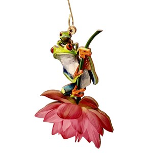 May include: A wooden ornament featuring two green tree frogs perched on a pink lotus flower. The larger frog is holding onto a green stem with its hind legs. The smaller frog is sitting on the larger frog's back. The ornament has a gold hanger.