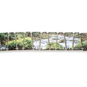 May include: A bracelet made of wood tiles with a nature scene printed on them. The tiles are connected by silver metal rings. The bracelet has a silver clasp.