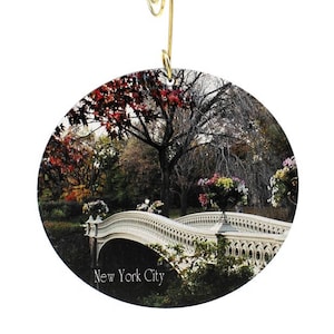 May include: A round, white, decorative ornament with a picture of a bridge in a park. The bridge is white with a stone arch design. The park is filled with trees, some with red and orange leaves. The text "New York City" is printed on the ornament.