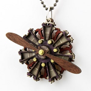 May include: A wooden pendant necklace featuring a detailed model of a radial engine with a propeller. The engine is painted in shades of purple, red, and white, with gold accents. The propeller is made of dark wood.