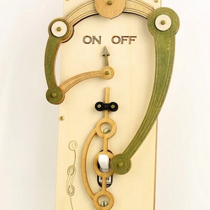 May include: A wooden light switch with a unique design featuring a lever and gears. The switch is labeled "ON OFF" and has a white toggle switch. The switch is made of light wood with green and black accents.