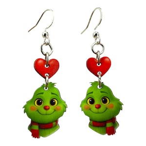 May include: A pair of dangle earrings featuring a green cartoon character with a red scarf and a red heart. The earrings have silver-colored hooks and are designed with a festive holiday theme.