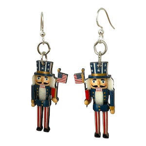 May include: A pair of patriotic earrings featuring Uncle Sam nutcracker figures. Each earring has a blue jacket, red and white striped pants, and a top hat with stars. They hold miniature American flags.