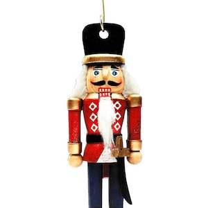 Puede incluir: Un adorno de cascanueces de madera con un uniforme rojo y blanco, un sombrero negro, una barba blanca y detalles dorados. El cascanueces sostiene una espada dorada y lleva un cinturón negro.