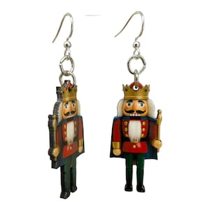 May include: A pair of silver-tone dangle earrings featuring wooden nutcracker soldier figures. The nutcrackers are dressed in red jackets, blue capes, green pants, and gold crowns. The earrings are designed with silver-tone fish hook ear wires.
