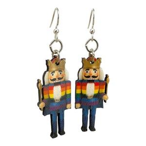 May include: A pair of wooden Nutcracker earrings. Each earring features a colorful rainbow-striped torso, a gold crown, and a white mustache. The earrings hang from silver-colored fish hook ear wires.