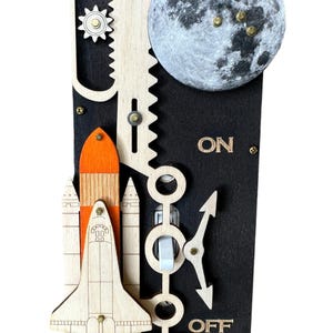 May include: A wooden light switch cover with a space theme. It features a moon, a rocket ship, and a toggle switch with "ON" and "OFF" labels. The cover is painted black with wood accents.