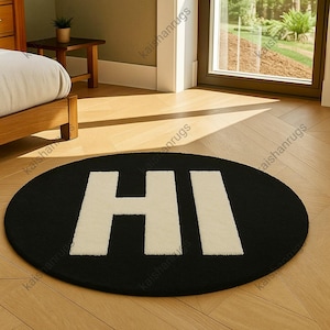 May include: Black oval rug featuring the word "HI" in large white letters. The rug is placed on a light-coloured wooden floor. The rug is a decorative item for a home.