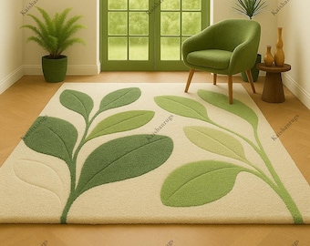 May include: A rectangular area rug with a cream-colored background and a large, stylized leaf design in shades of green. The leaves are textured and create a 3D effect. The rug is placed on a wooden floor, with a green chair and potted plant in the background.