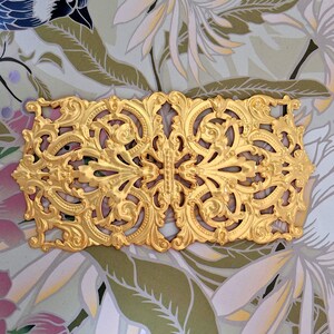 May include: A rectangular gold-colored decorative applique featuring an intricate, symmetrical scrollwork pattern with raised relief details. The piece has a metallic finish and a complex, ornate design with swirling foliage motifs throughout its surface.