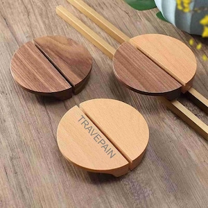 Op de afbeelding: Set van drie houten eetstokjessteunen. Elke steun is rond en in twee helften verdeeld. Twee zijn donkerbruin en één is lichtbruin met het woord "TRAVEPAIN" erop gedrukt. Eetstokjes zijn ook zichtbaar.