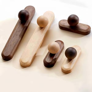 Może przedstawiać: Kolekcja drewnianych gałek do szafek w różnych rozmiarach i wykończeniach. Gałki mają zaokrąglony kształt kuli na prostokątnej podstawie. Odcienie drewna wahają się od jasnego naturalnego do ciemnobrązowego. Do mebli i dekoracji wnętrz.