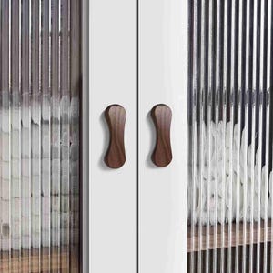 Könnte beinhalten: Nahaufnahme eines weißen Schranks mit zwei Holzgriffen. Die Schranktüren haben eine strukturierte Glasscheibe. Die dunkelbraunen Griffe haben ein geschwungenes Design. Die Glasscheibe hat vertikale Linien.