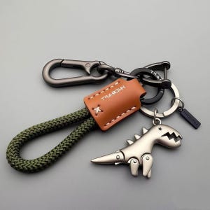 May include: A keychain with a silver dinosaur charm, a brown leather tag, and a green rope loop. The keychain has a metal clasp and a black ring. The leather tag has the word "HEBVEL" printed on it.