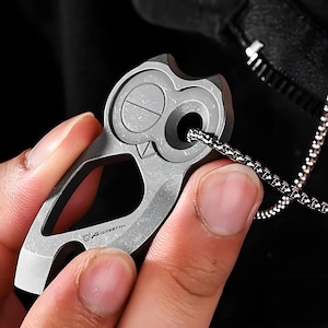 Pode incluir: Um pingente prateado em forma de coruja com uma corrente. O pingente tem um design recortado e um detalhe circular de olho. A corrente é feita de pequenas peças de metal ligadas. O pingente é segurado por uma pessoa.