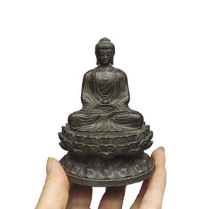 May include: A dark bronze-coloured Buddha statue in a seated meditation pose, resting on a detailed lotus flower base. The figure features intricate robe textures and a small swastika symbol on the chest, crafted with a weathered metallic finish.
