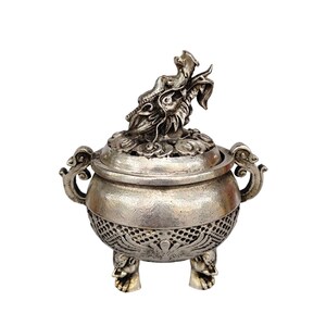 May include: A silver-colored incense burner with a dragon-shaped lid handle. The burner has a round body with decorative handles and rests on three ornate feet. The lid features a floral design, and the body has a patterned lower section.