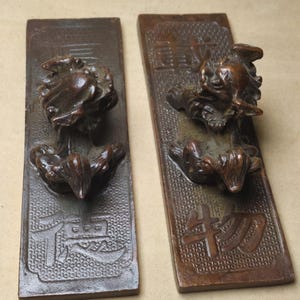 May include: A pair of antique bronze door pulls. Each rectangular plate features a detailed relief of a mythical creature with a textured background and Chinese characters. The door pulls are a dark brown colour.