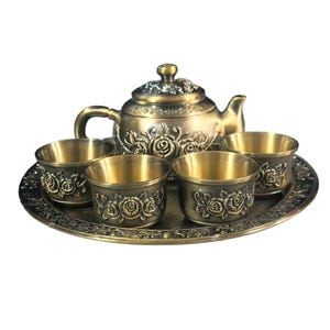 Pode incluir: Um conjunto de chá em metal com tom de bronze, composto por um bule e quatro chávenas a condizer dispostas num tabuleiro circular. Cada peça é decorada com motivos de rosas em relevo e padrões florais, com um acabamento antigo uniforme.