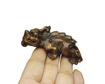 May include: A bronze-colored figurine of a mythical creature, possibly a dragon or lion, with intricate details. The sculpture is held in a hand against a white background. The figurine has a textured surface and a detailed mane.