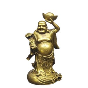 Puede incluir: Una figura de metal de color dorado de un personaje sonriente y corpulento de pie sobre una base. El personaje sostiene un lingote de oro en alto con una mano y un cetro ruyi en la otra. La escultura presenta túnicas fluidas y detalladas.