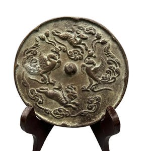 May include: A circular bronze-toned metal mirror featuring a raised relief design of two birds and two four-legged creatures surrounding a central knob. The surface has a weathered, textured finish with swirling cloud-like patterns throughout.