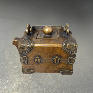 May include: A bronze-colored, box-shaped teapot with ornate detailing. The teapot features a spout, a lid with a spherical knob, and decorative elements on the corners and sides. It has two handles and rests on four small feet.
