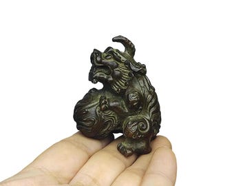 May include: A bronze-colored, detailed figurine of a mythical creature, possibly a guardian lion. The sculpture has intricate carvings and a textured surface. The creature is depicted in a seated position, holding a ball-shaped object.