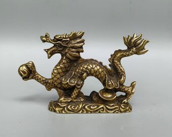 Puede incluir: Figurilla de dragón de latón detallada con escamas intrincadas, una melena fluida y una bola sostenida en su garra. El dragón está posado sobre una base decorativa, mostrando un diseño tradicional asiático. La figura tiene un tono dorado.
