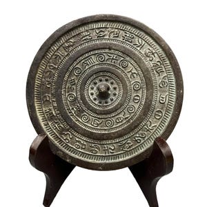 May include: A circular dark grey metal mirror featuring a central knob surrounded by concentric rings of embossed spiral patterns and a wide outer border filled with intricate, repeating calligraphic characters in a weathered, aged finish.