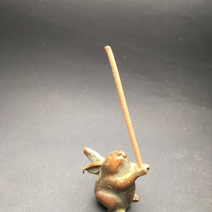 May include: A bronze rabbit figurine holding a brown incense stick. The rabbit is depicted in a standing pose with its ears raised. The figurine has a detailed texture and is set against a dark background.