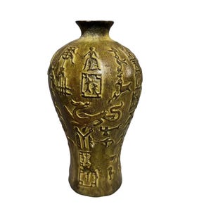 May include: A tall, bulbous vase with a textured, metallic gold finish. The surface is decorated with embossed, dark-toned relief carvings featuring various stylized figures, animals, and geometric symbols arranged in a vertical, repeating pattern.
