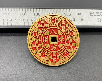 May include: A round, red and gold coin with Chinese characters and symbols. The coin has a square hole in the center and is approximately 2 inches in diameter. The coin is placed next to a ruler.