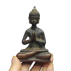 May include: A small, dark bronze-toned metal figurine of a seated Buddha in a meditative pose with hands pressed together in front of the chest. The figure features a textured, pointed ushnisha on the head and draped robes over the shoulders.