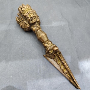 May include: A bronze ritual dagger with a detailed handle depicting a multi-faced deity and intricate patterns. The blade tapers to a point, with a serpent design along the edges. The dagger is likely of cultural or religious significance.