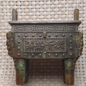 May include: A bronze-colored, three-legged incense burner with a rectangular body and two handles. The front of the burner features an inscription in Chinese characters. The burner has decorative patterns and a weathered appearance.