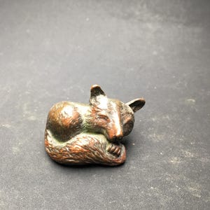 May include: A small, bronze-colored sculpture of a curled-up fox. The fox is in a sleeping position, with its paws tucked in and its tail wrapped around its body. The sculpture has a detailed texture and a naturalistic appearance.