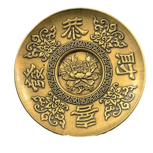 Puede incluir: Un plato circular de color latón con una peonía en relieve en el centro, rodeada por un borde geométrico. Cuatro caracteres chinos están dispuestos alrededor del centro, intercalados con cuatro motivos florales detallados.